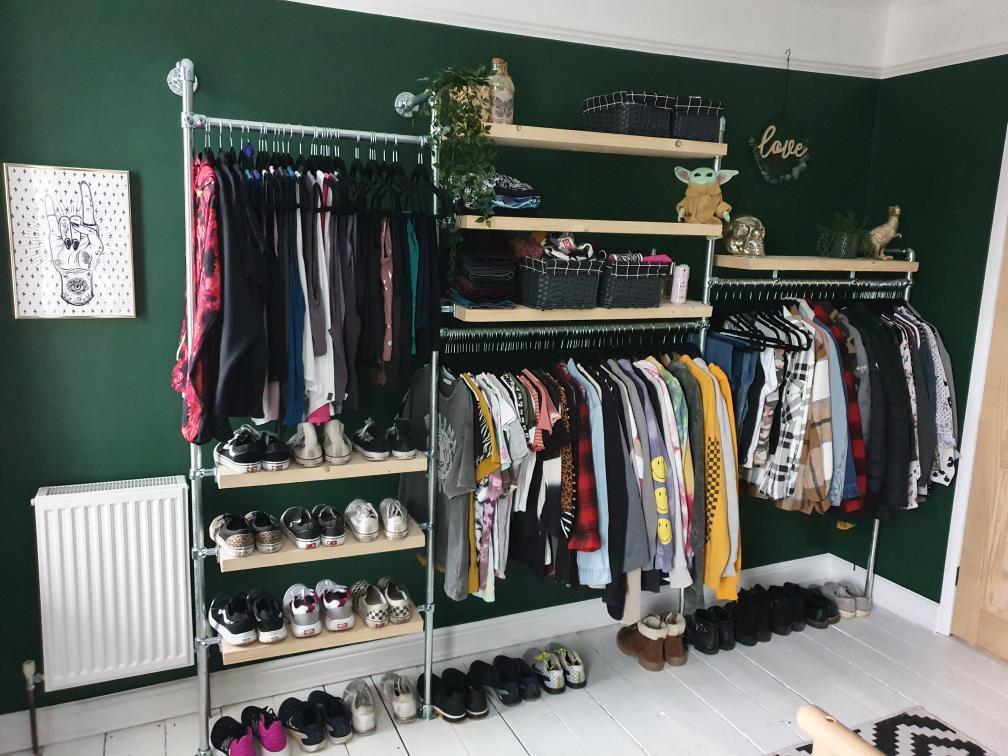 A multi-level bedroom tube clamp wardrobe area, sat against a deep green wall, complete with scaffold board shelves, house plants and baskets for decoration.