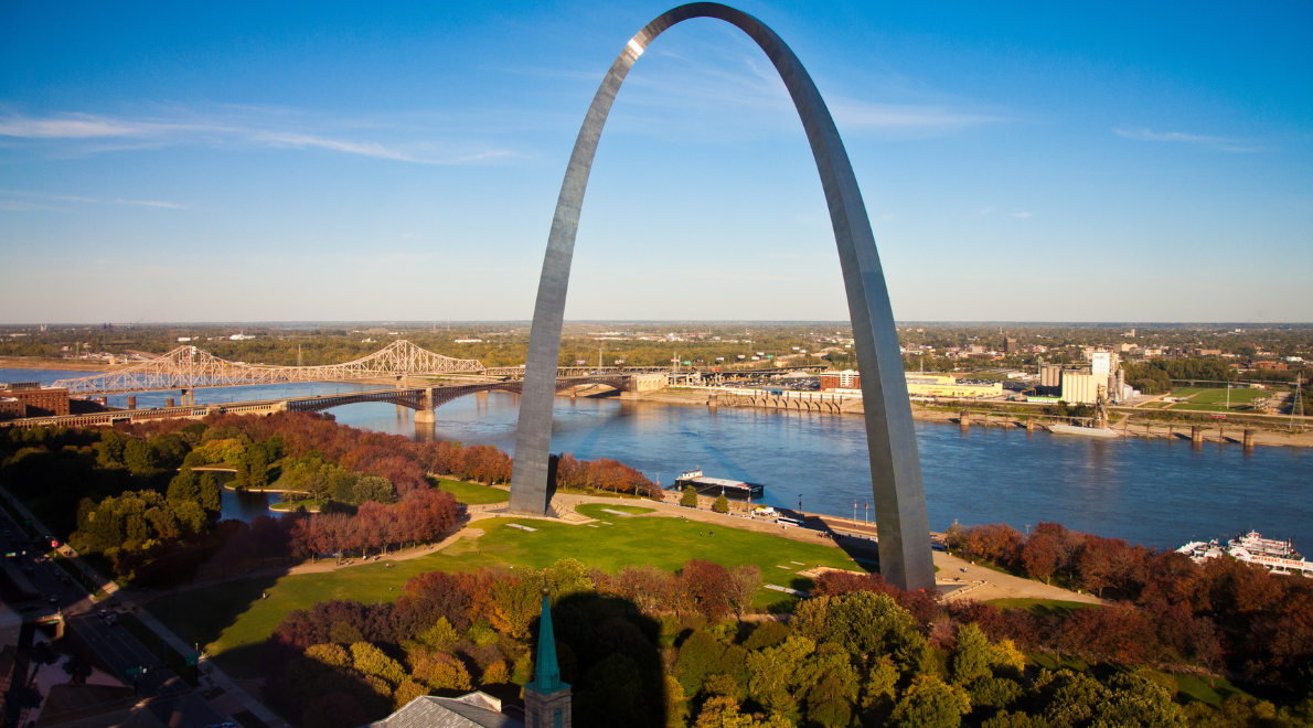 Scenic panorama of the Gateway Arch framed by lush greenery and the flowing river - Nature and architecture in harmonious embrace. The stainless steel arch dominates the skyline, while the tranquil river and distant bridge create a picturesque contrast, illustrating the intersection of human achievement and the beauty of the natural world