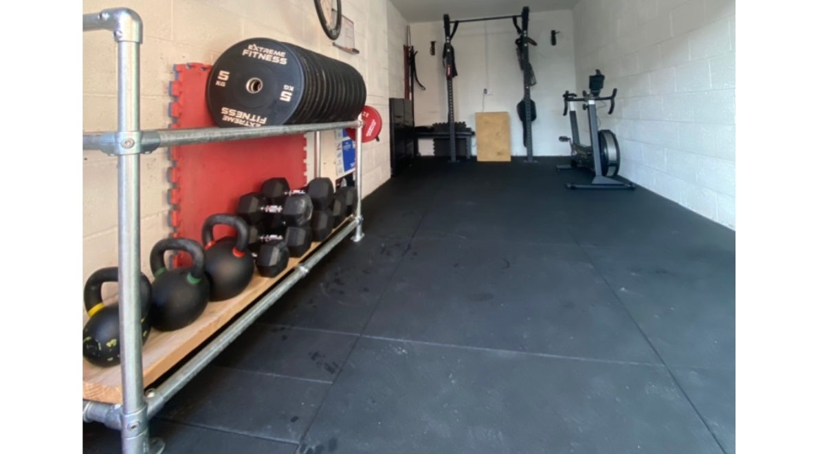 Home garage gym complete with a white breeze block walls, cushioned floor, exercise bike, pull-up station and a DIY weight rack made from galvanised tube and clamp fittings to house weight plates, kettlebells and hex dumbbells