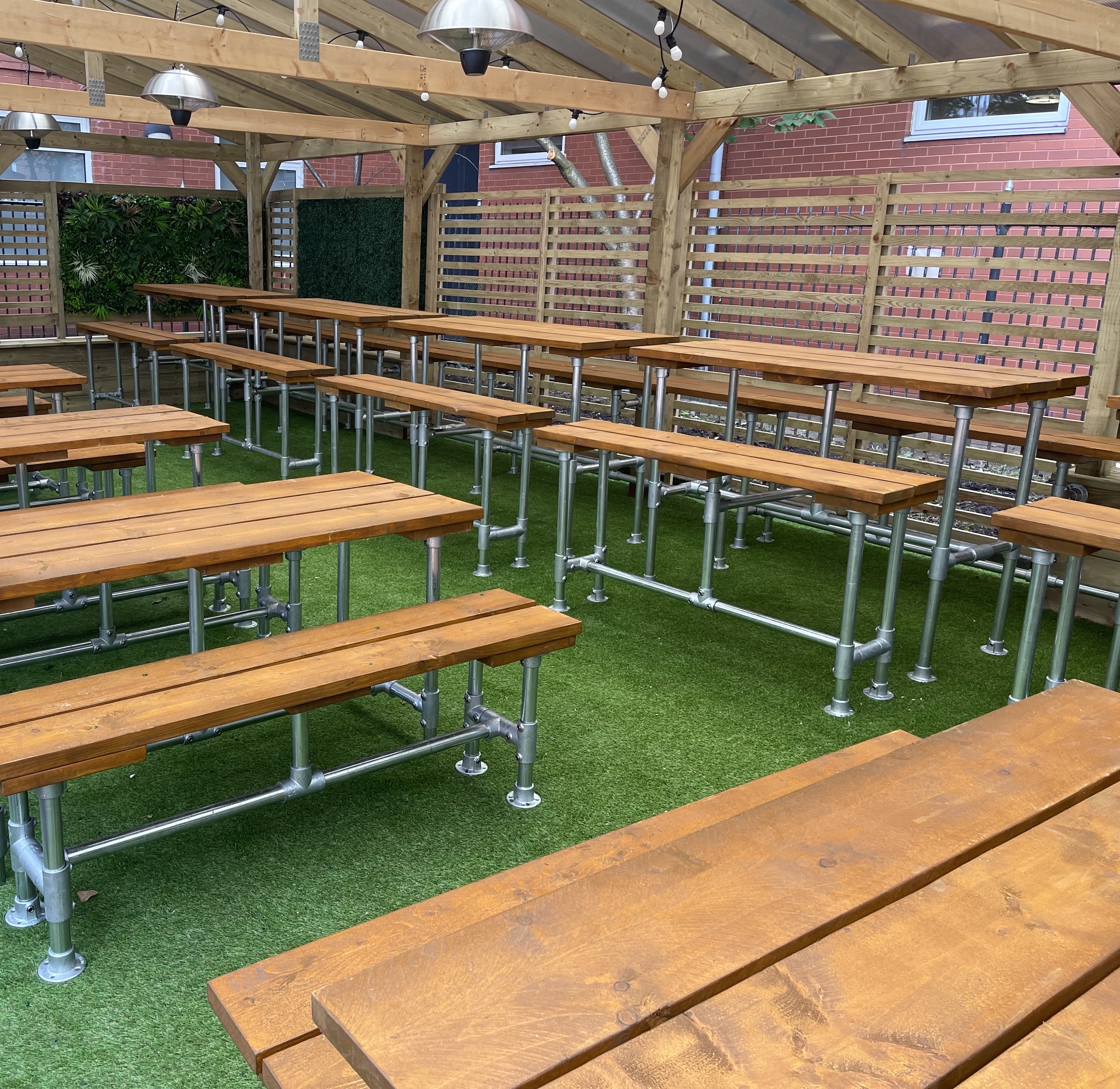 An outdoor seating area made up of a timber overhead structure, artificial grass floor and a number of tube and clamp picnic tables and benches with treated scaffold board tops