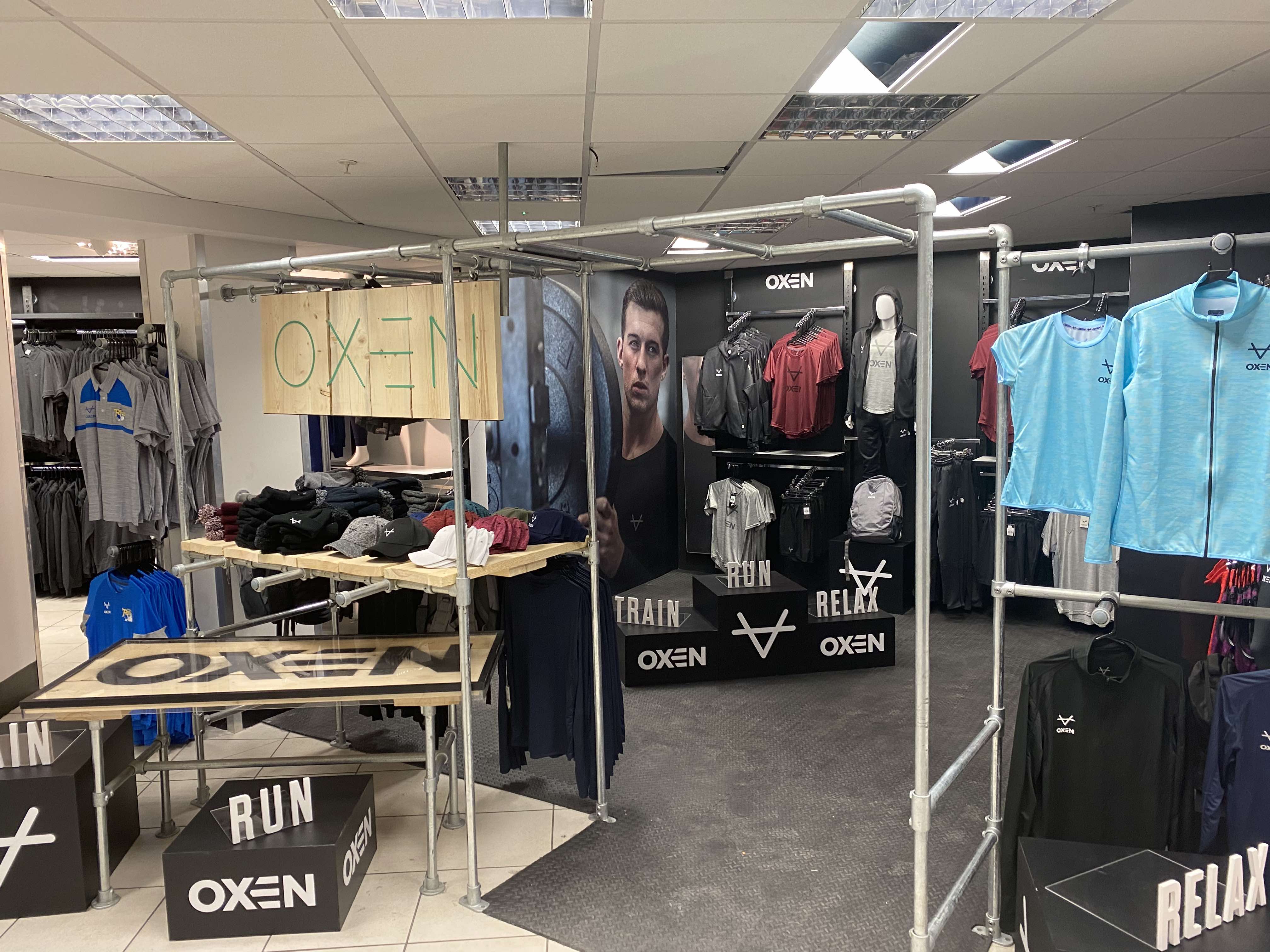 A sports clothing section of a retail store with matte black walls, steel treadplate flooring and a galvanised steel scaffolding frame to create an entrance to this area of the store and scaffolding boards to create signage and shelving to display clothes>
  </div>
  <div class=