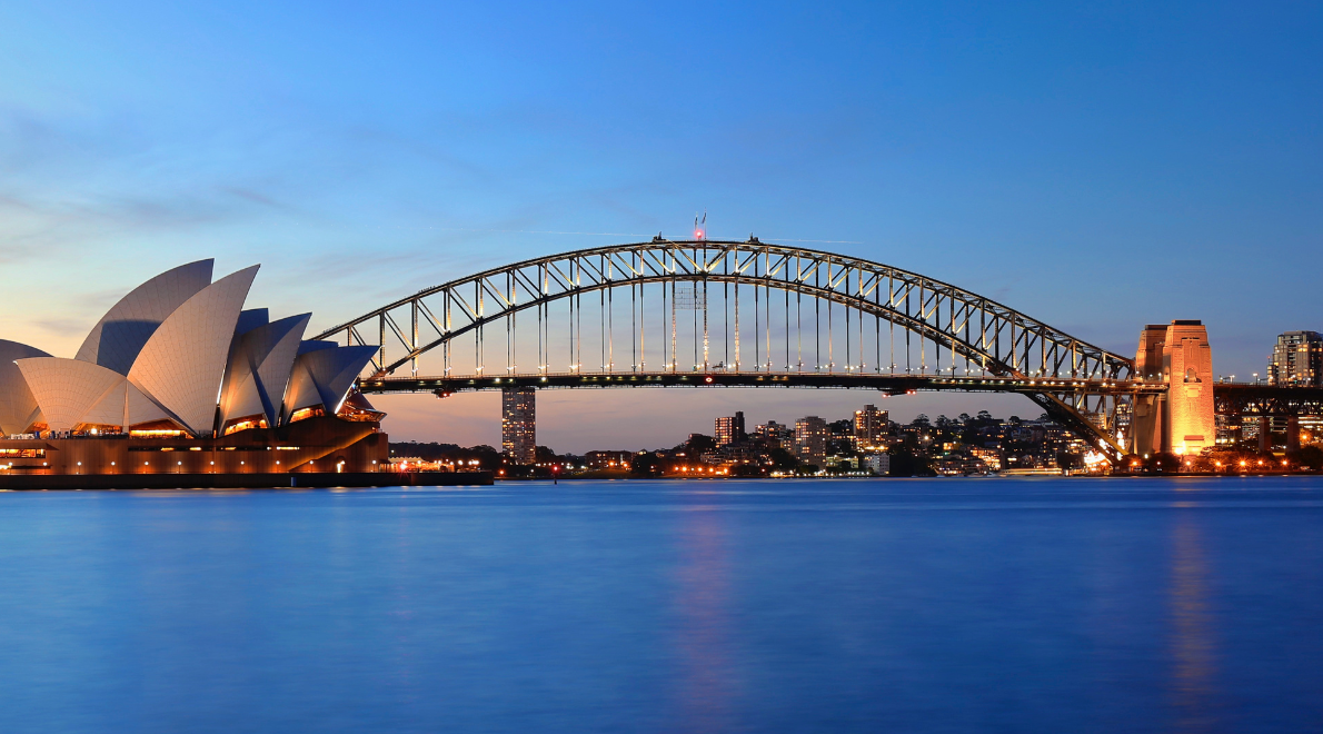 Tranquil waterfront view framing the Sydney Harbour Bridge - An engineering marvel gracefully arching over shimmering waters. The iconic steel structure connects two shores, its graceful lines mirrored in the gentle ripples of the harbor. To the left, the Sydney Opera House stands as a sculptural masterpiece, forming a harmonious duo that symbolizes Sydney's architectural and cultural prominence.