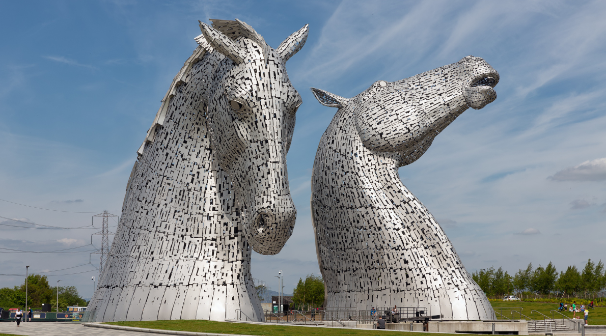 Close-up encounter with The Kelpies - Monumental equine sculptures rise before you, their shimmering metallic skin reflecting the sunlight. The intricate details of the steel horses come to life as you stand at their feet, feeling the sense of mythical power and fluidity captured in these towering works of art.