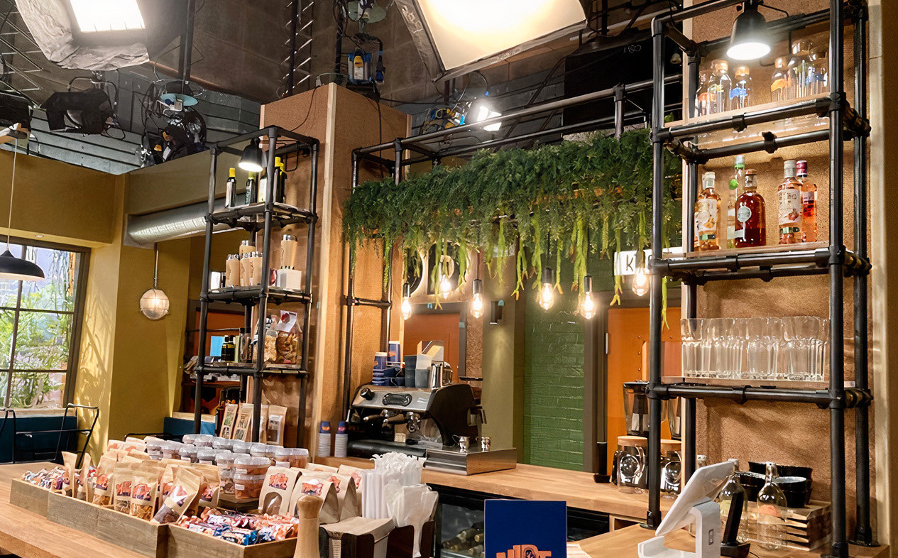 Looking over the counter of a warm, modern coffee shop with wooden worktops, green tiled walls, low hanging lightbulbs and industiral black tube and clamp shelving used to display glasses, baked gods and bottled drinks